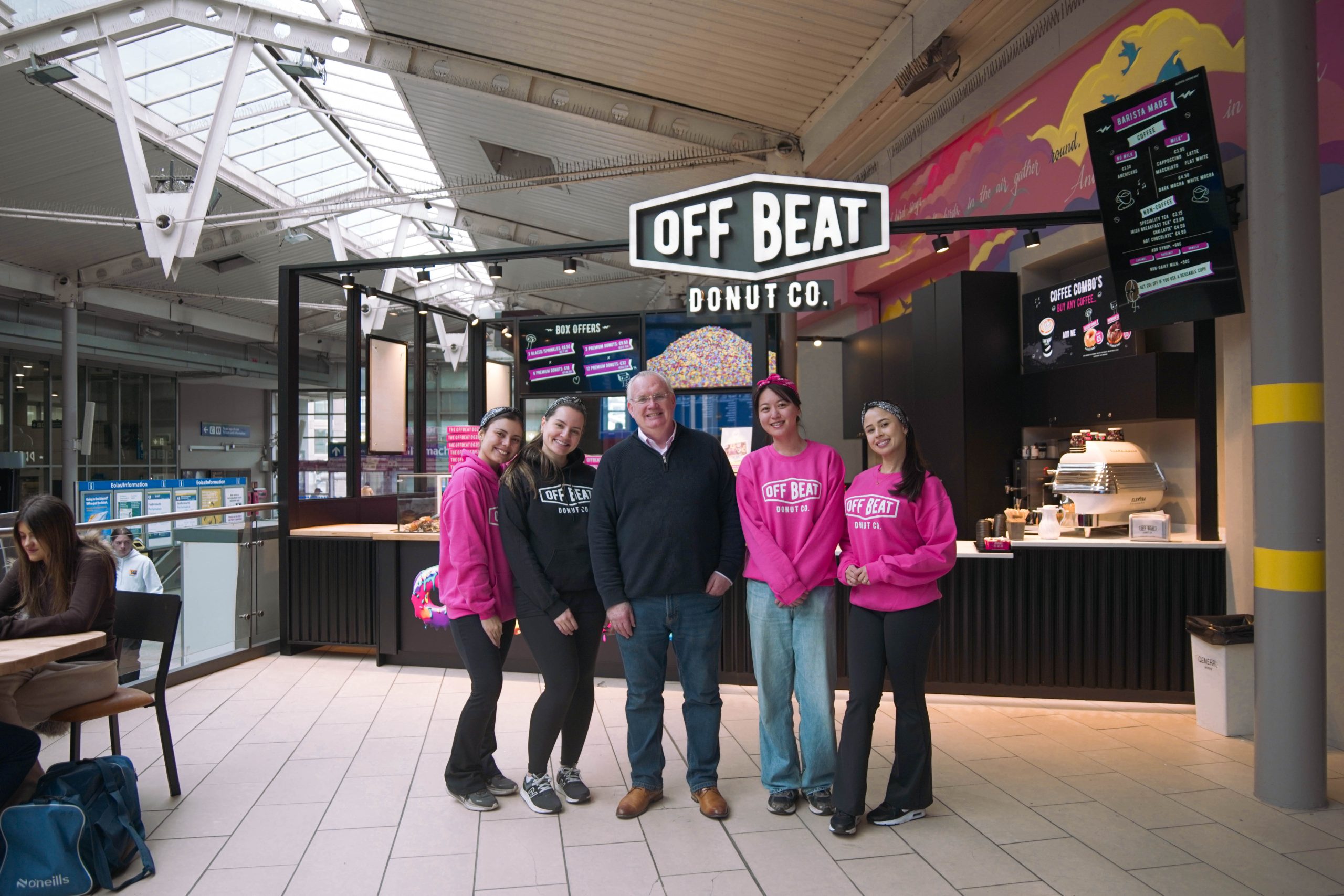 Offbeat Donuts founder Brian stands smiling alongside team members in pink branded hoodies in front of the new Connolly Station store, with the donut counter and menu boards behind them.