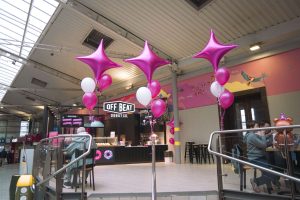 Off Beat Donut Co. kiosk decorated with pink balloons at Connolly Station in Dublin, with customers seated nearby in a bright indoor train station setting.